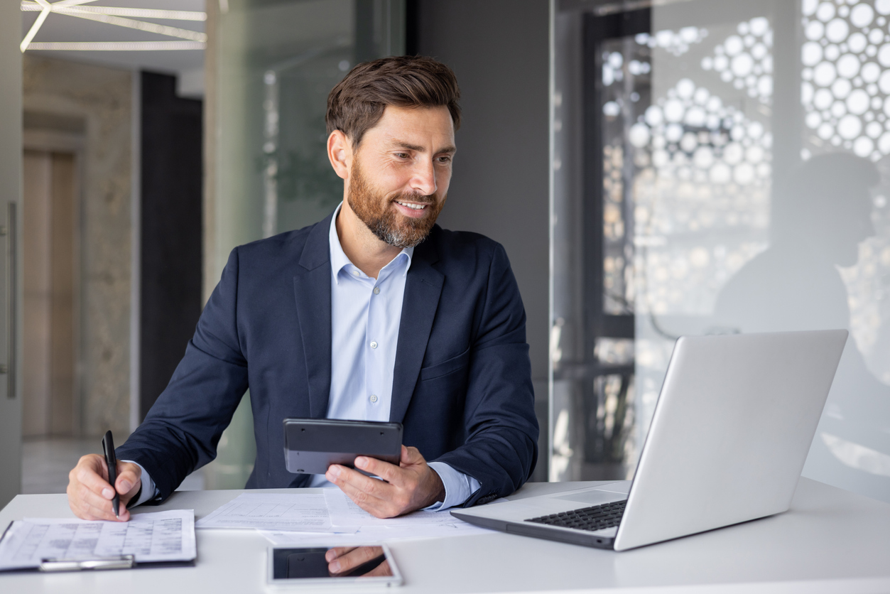 Smiling young man financial expert, businessman sitting in the office at the desk, working on a laptop and with documents, using a calculator